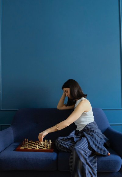 Tablero de ajedrez con piezas oscuras y claras, marco de madera, sobre un sofá de tela azul. Persona vestida con una blusa blanca y pantalones a rayas alcanza una pieza.