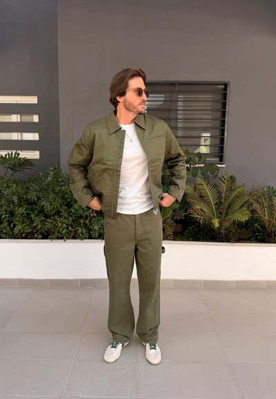 Hombre con gafas de sol, chaqueta y pantalones verde oliva, zapatillas blancas y camiseta, de pie con las manos en las caderas cerca de plantas y una ventana.