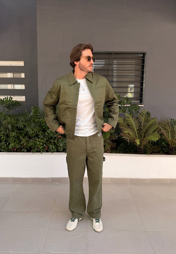 Mees päikeseprillide, oliivrohelise jope ja pükstega, valgete tossude ja t-särgiga, seisab käed puusas taimed ja aken lähedal.