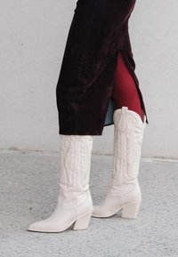 Białe kowbojskie buty ze skóry z zaostrzonym noskiem i szerokim obcasem, zdobione wytłaczanymi wzorami na cholewce, noszone z ciemną welwetową spódnicą.