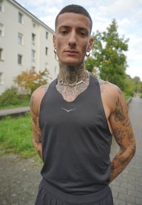 Mladý tatovaný muž s krátkymi vlasmi a piercovaným nosom, oblečený v čiernom tielku a reťazovom náhrdelníku, stojí vonku pri budovách a zeleňi.