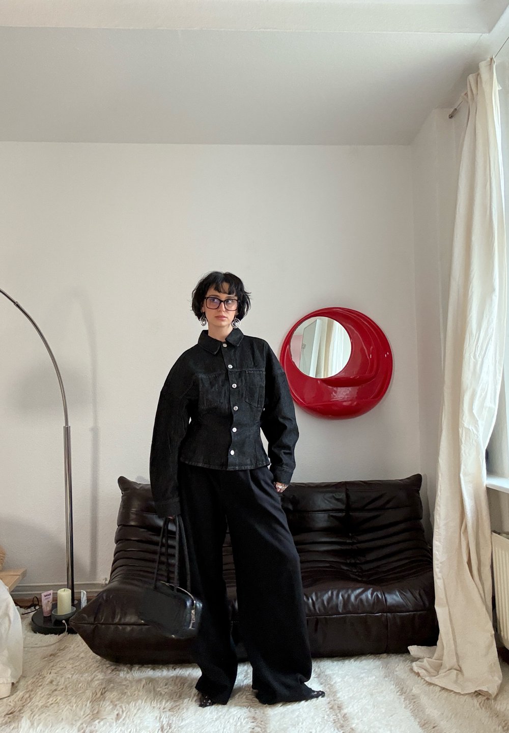 Personne aux cheveux courts foncés et portant des lunettes, vêtue d'une veste noire et d'un pantalon large, se tient dans un salon avec un canapé brun, un miroir rond rouge et des rideaux blancs.