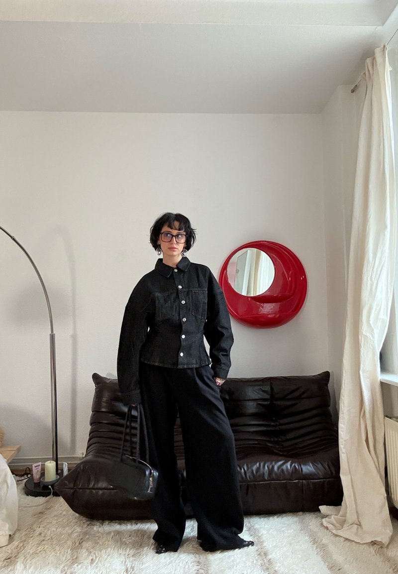 Personne aux cheveux courts et foncés portant des lunettes, une veste noire et un pantalon large, se tenant dans un salon avec un canapé marron, un miroir rond rouge et des rideaux blancs.