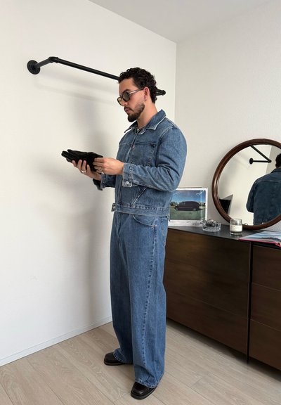 Hombre con chaqueta vaquera y jeans sosteniendo guantes negros doblados junto a una cómoda con un espejo redondo y un marco de foto.