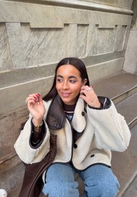 Jeune femme assise sur des marches extérieures, portant une veste en shearling crème et un jean bleu, ajustant ses cheveux avec un léger sourire.