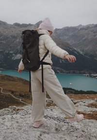 Čierny turistický batoh s textúrovaným vzorom, nosený cez bielu zateplenú bundu, v kombinácii s béžovými nohavicami a ružovými teniskami, na pozadí hôr.