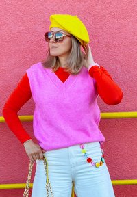 Roze wollen vest over een rode lange mouwen top, gecombineerd met witte jeans. Gele baret en oversized zonnebril. Multicolore kettingaccessoire rond de taille.