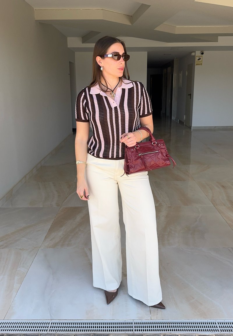 Femme portant un polo rayé, un pantalon blanc à jambes larges, des talons pointus marron, des lunettes de soleil, tenant un sac à main bordeaux, debout dans un couloir.