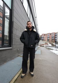 Homme portant une veste noire avec col en fourrure, pantalon noir et chaussures beiges, debout sur un trottoir urbain à côté d'un bâtiment moderne. Neige visible au sol.