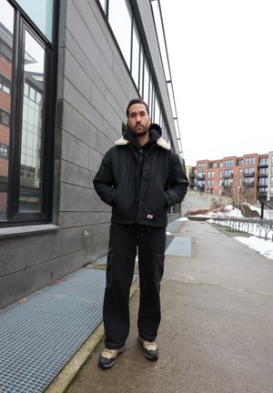 Hombre vestido con chaqueta negra con cuello de piel, pantalones negros y zapatos beige, de pie en una acera urbana junto a un edificio moderno. Se ve nieve en el suelo.