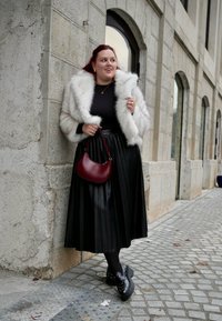 Λευκό μπουφάν από faux γούνα, μαύρη πλισέ φούστα, μαύρες καλσόν και μαύρα παπούτσια με ανάγλυφα. Κρατάει μια μικρή κόκκινη τσάντα και στέκεται μπροστά σε έναν πέτρινο τοίχο.