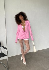 Pink tailored blazer over a white top, paired with lace patterned tights. White pointed-toe heels and a small white handbag.