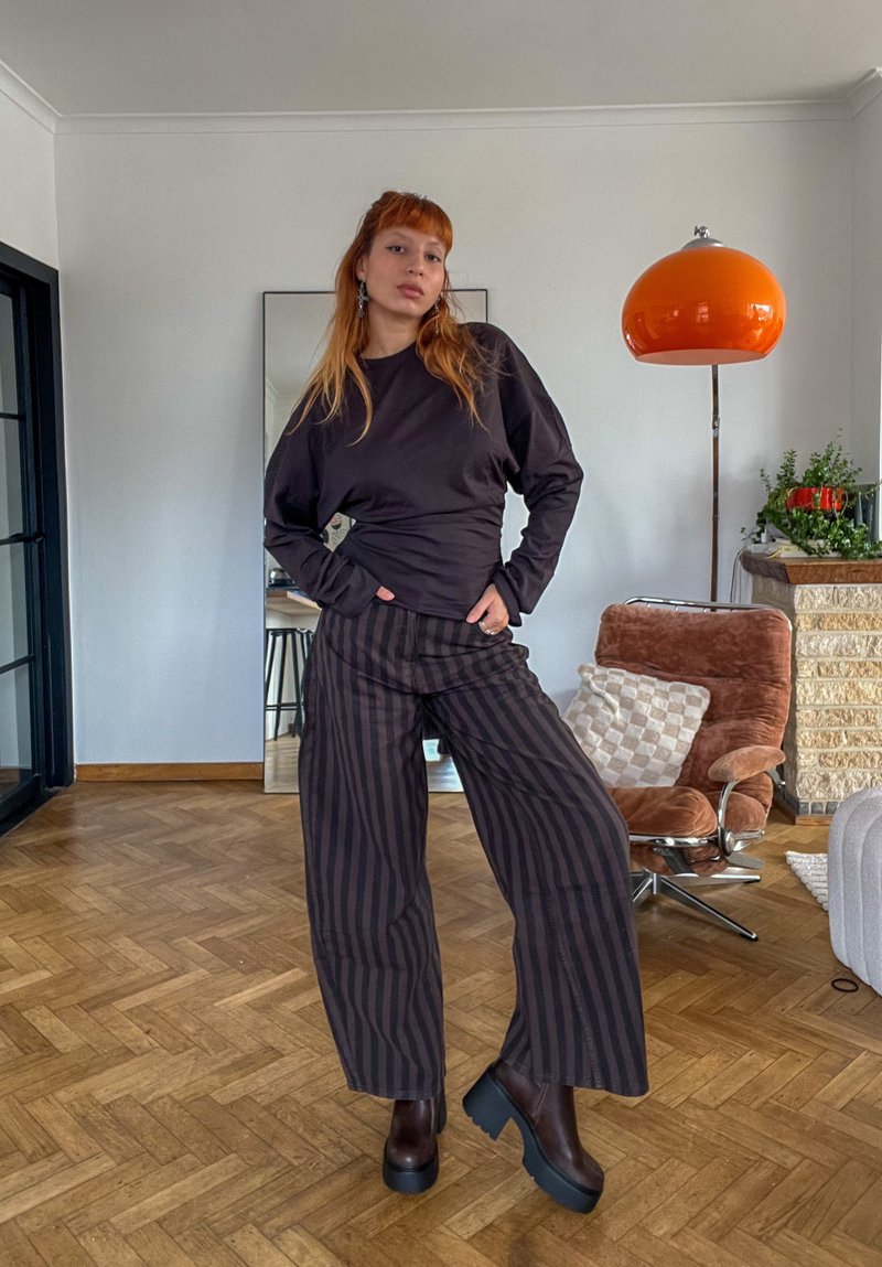 Jeune personne aux longs cheveux roux porte un haut noir et un pantalon large rayé foncé, debout dans un salon moderne avec un sol en bois.