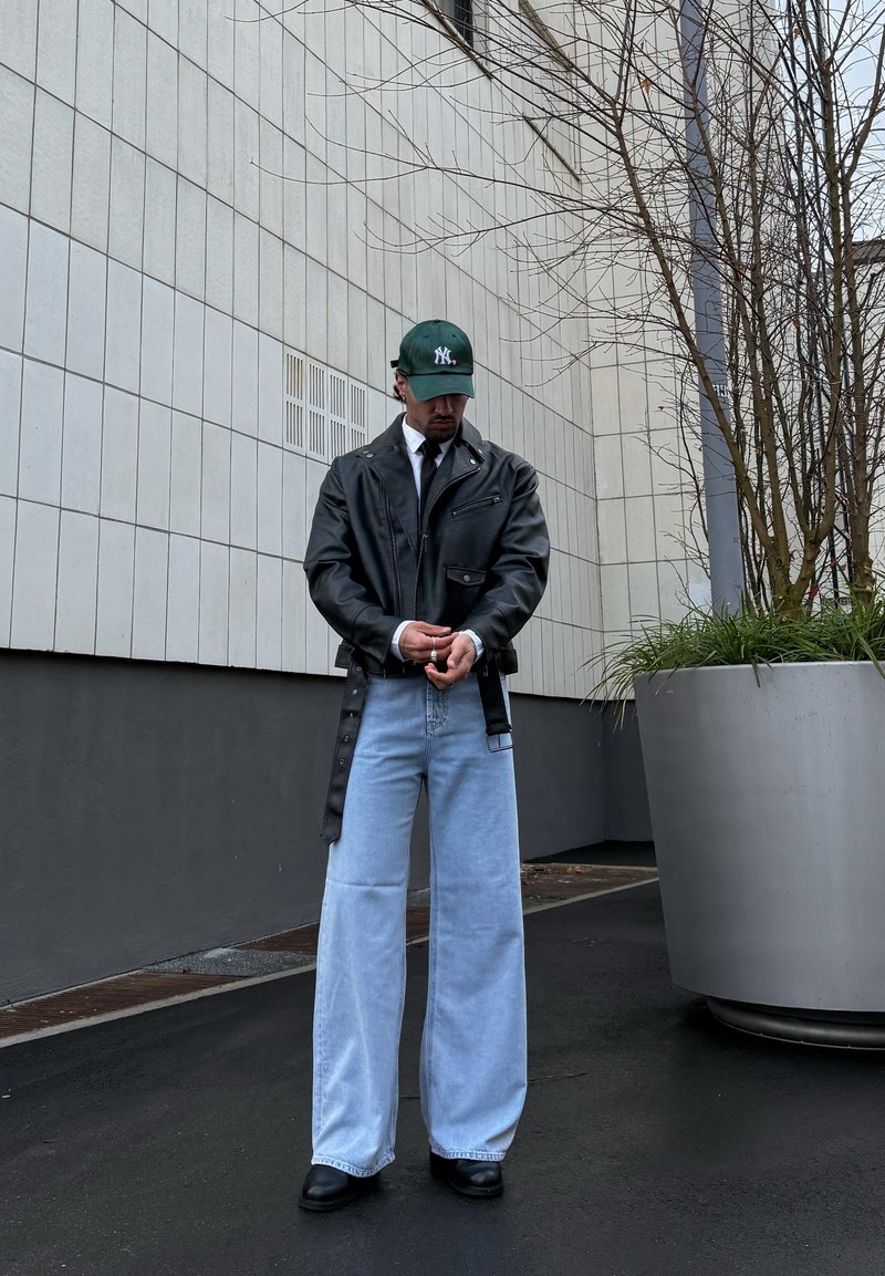 Hombre con chaqueta de cuero negra, vaqueros de pierna ancha azul claro, zapatos negros y una gorra de béisbol verde, de pie cerca de una maceta grande al aire libre.