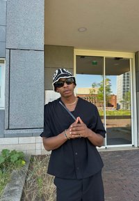 Maglietta nera a maniche corte e pantaloni abbinati, accessoriati con un cappello a secchiello con motivo bianco e nero e occhiali da sole, in piedi contro un muro di pietra.