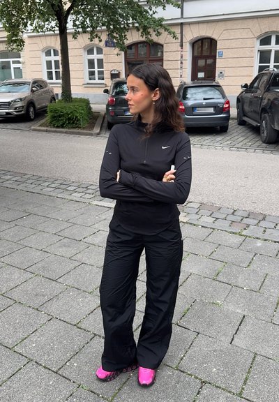 Sudadera negra de manga larga con cremallera y pantalones sueltos a juego. Presenta un logo blanco y zapatillas rosas con suelas texturizadas, todo sobre un fondo de adoquines.