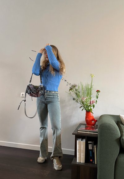 Mujer con cabello rubio largo, vestida con un suéter azul y jeans, sostiene un bolso floral, de pie junto a una mesa con flores en un jarrón rojo.