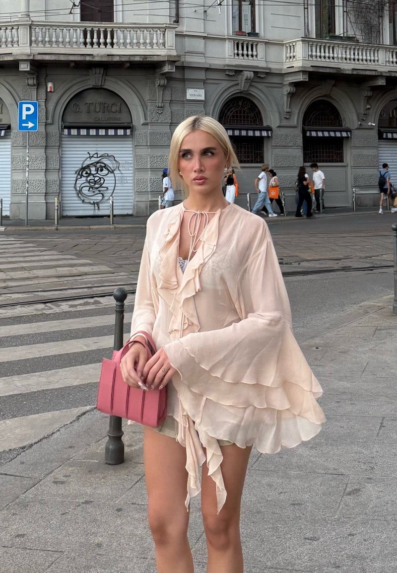 Blusa de un rosa claro, con volante y mangas largas acampanadas, y cuello con cordón. Sosteniendo un pequeño bolso estructurado rosa con acentos verticales.