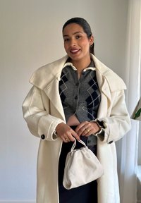 Fehér gyapjú kabát szürke, kockás mintájú, rövid fazonú pulóveren. Egy kis krémszínű textil táskát tart a kezében. Egyszerű gyűrűk kiegészítőként.