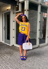 Purple Lakers jersey with the number 23, teamed with purple shorts. White handbag featuring a multicoloured logo, blue shoes, and a purple headband.
