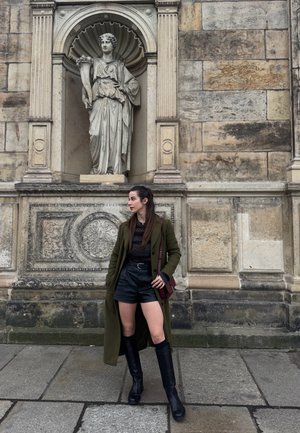Mujer con abrigo verde y botas negras posando sobre pavimento de piedra frente a una pared de piedra ornamentada con una estatua clásica en un nicho superior.