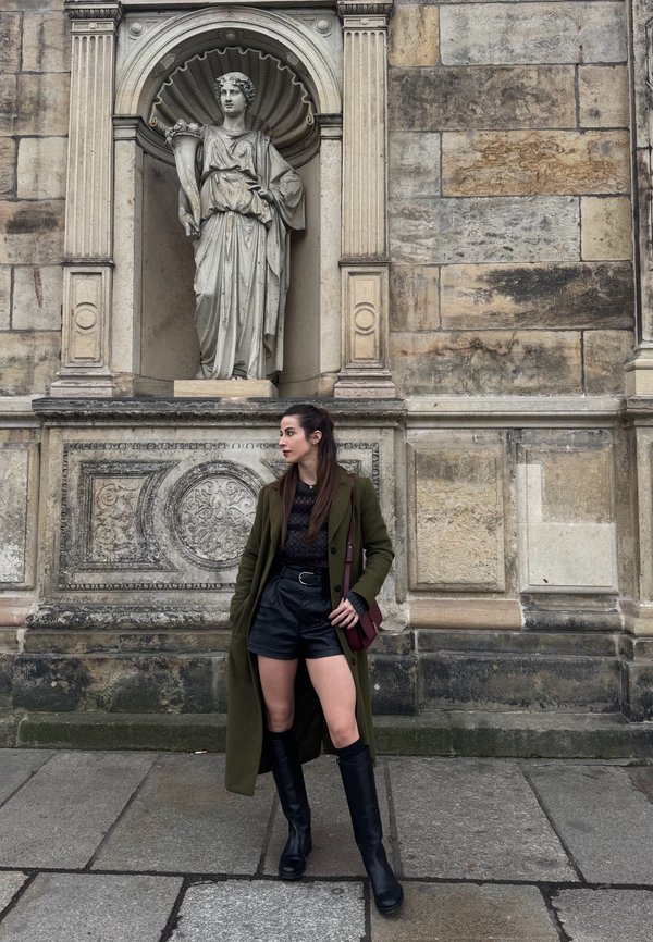Mujer con abrigo verde y botas negras posando sobre pavimento de piedra frente a una pared de piedra ornamentada con una estatua clásica en un nicho superior.