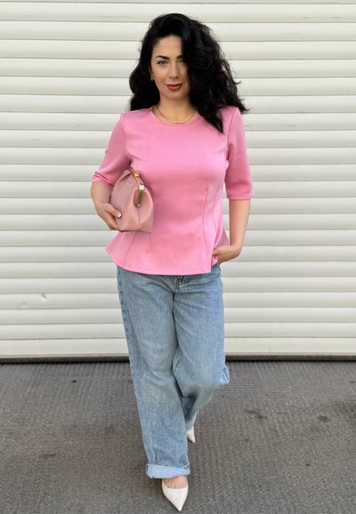 Blusa rosa de manga tres cuartos con costuras laterales, combinada con jeans de corte relajado en azul claro y tacones blancos de punta. Sosteniendo un bolso de mano rosa claro.