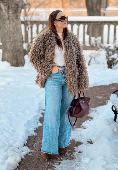 Abrigo marrón de pelo sintético, top blanco acanalado, vaqueros anchos azul claro, botas marrones de tacón y bolso de mano color púrpura oscuro, sobre un fondo de nieve.