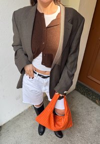Grå mønstret blazer over en brun cardigan, hvid kortærmet skjorte, hvide shorts, knæhøje sorte støvler og en orange vævet håndtaske.