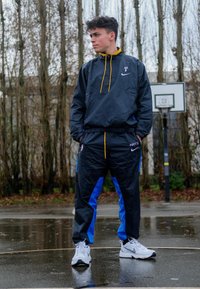 Fato de treino preto em nylon com painéis laterais azuis e detalhes em zíper amarelo. Ténis brancos da Nike. O fundo apresenta uma tabela de basquetebol e árvores despidas.