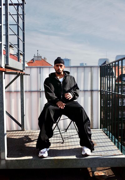 Sudadera negra con cremallera, pantalones anchos negros y zapatillas blancas. Sentado en una silla plegable sobre una plataforma de metal con un fondo de paisaje urbano.