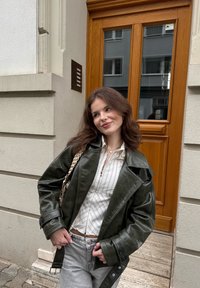 Zielona skórzana kurtka z dużymi klapami, w połączeniu z białą koszulą w paski i jasnoszarymi dżinsami. W tle drewniane drzwi.