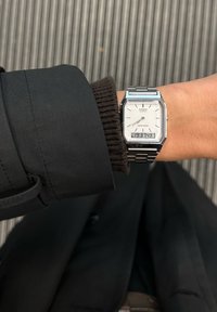 Strieborné hodinky Casio na quartz s osemuholníkovým ciferníkom, bielym displejom, zobrazením dátumu a kovovým náramkom, nosené na zápästí zakrytom čiernym rukávom kabáta.