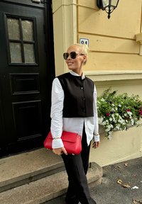 Μαύρο κοντό γιλέκο πάνω από ένα λευκό πουκάμισο με κουμπιά, συνδυασμένο με μαύρο παντελόνι. Κρατάει μια κόκκινη δερμάτινη τσάντα. Μαύρα γυαλιά ηλίου. Πράσινα φυτά στο φόντο.