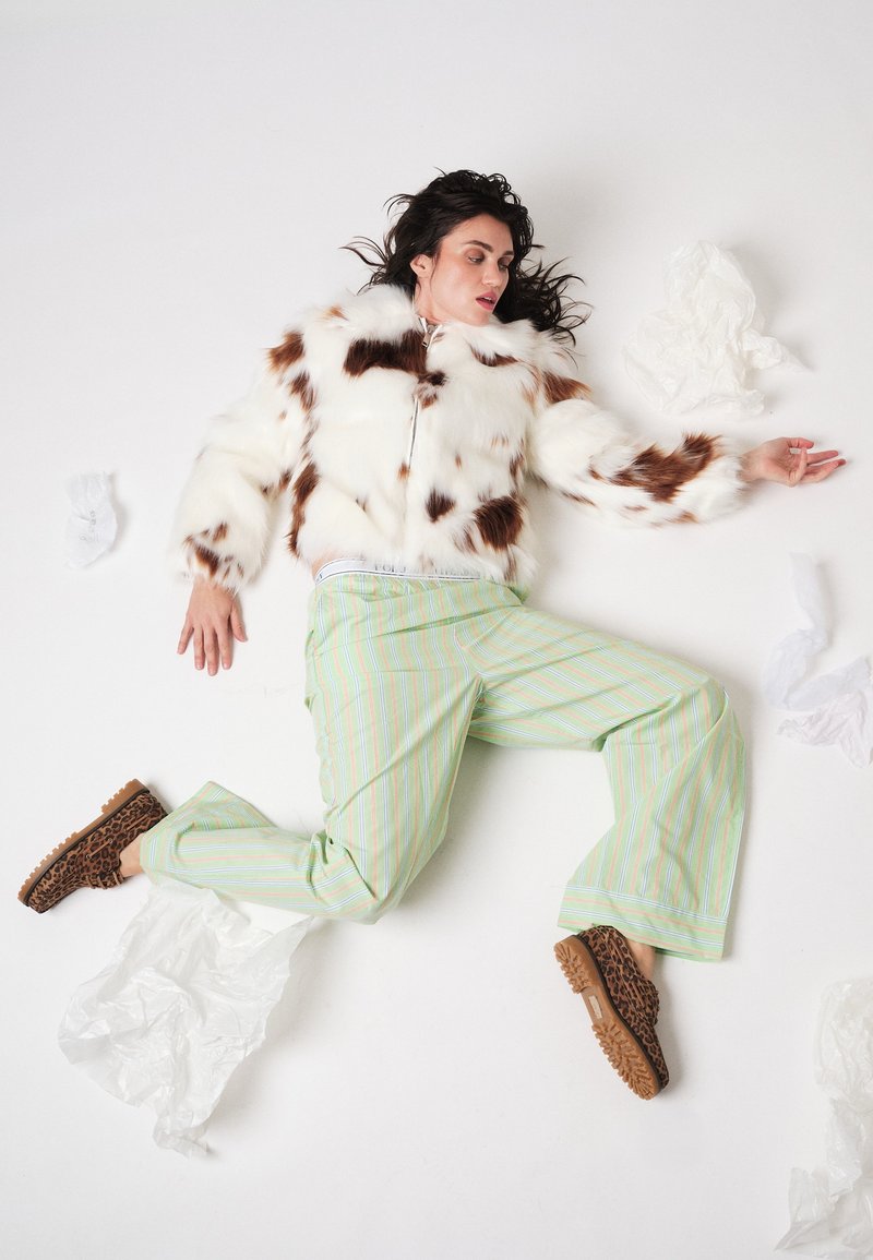 Vrouw liggend op een witte vloer met een gevlekte bontjas, groene gestreepte wijde broek, schoenen met luipaardprint, omringd door gekreukeld wit plastic.