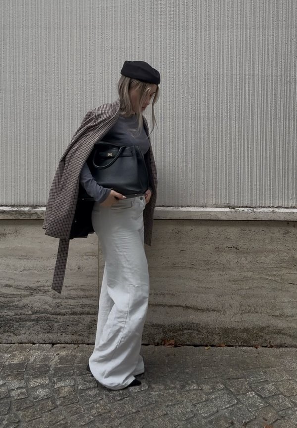 Mujer con pantalones blancos de pierna ancha, camiseta gris de manga larga, chaqueta a cuadros, boina negra, llevando bolso negro, caminando junto a una pared con textura.