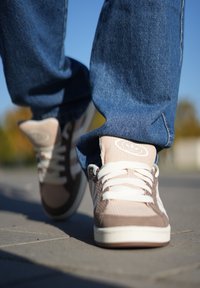 Brune og lyserøde sneakers med tekstureret stof, hvide snørebånd og logo på tungen. Jeans dækker lidt over toppen af skoene.