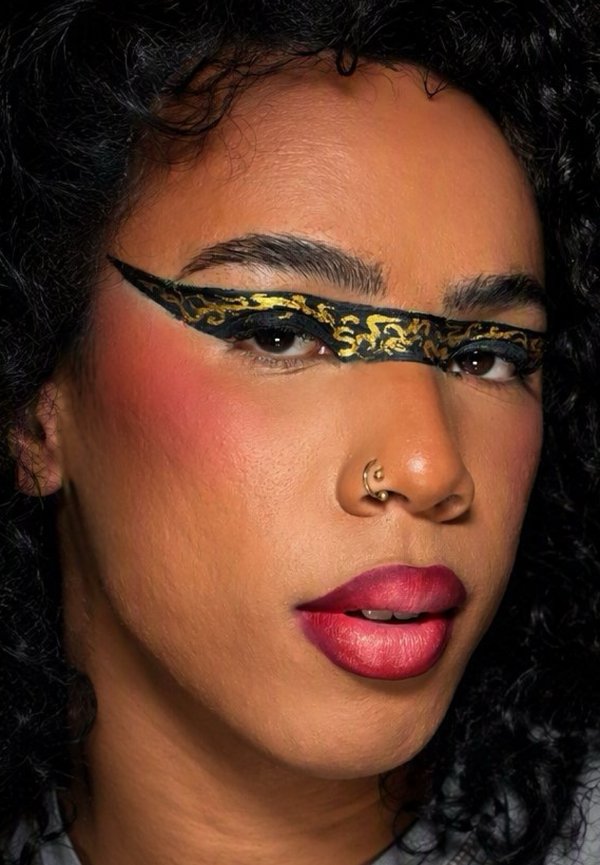 Persoon met krullend haar en een goud-zwart patroon oogmasker-make-up, opvallende rode lippenstift, blush en een dubbele neusring in het linker neusgat.
