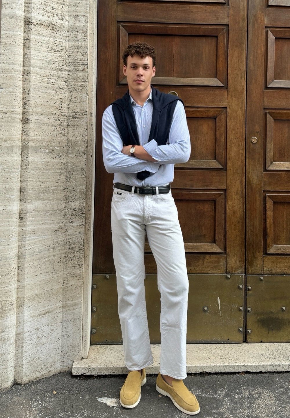 Ανοιχτό μπλε καρό πουκάμισο, λευκά παντελόνια, μπεζ μπαρέτες. Ναυτικό πουλόβερ ριγμένο στους ώμους. Φόντο ξύρινης πόρτας.