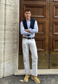 Ανοιχτό μπλε καρό πουκάμισο, λευκά παντελόνια, μπεζ μπαρέτες. Ναυτικό πουλόβερ ριγμένο στους ώμους. Φόντο ξύρινης πόρτας.