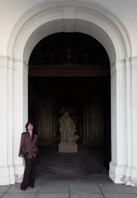 Personne en costume violet foncé appuyée contre une porte blanche en arc, avec une statue en pierre classique visible à l'intérieur d'un bâtiment faiblement éclairé.
