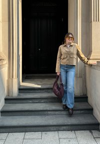 Beige lærjakke med kort lengde, hvit skjorte, lyseblå flagrende jeans, brune platåsko, og en mørk lilla vevd håndveske mot en stein bakgrunn.