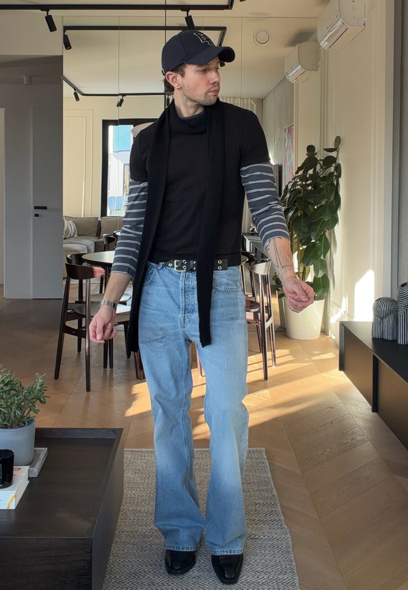 Homme portant une casquette noire, des hauts superposés noirs avec des manches rayées, un jean bleu clair, des chaussures noires, debout dans un salon moderne avec des plantes et des meubles.