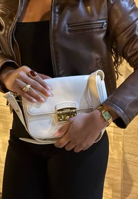 Borsa a tracolla bianca in pelle con una chiusura in metallo dorato, portata a mano. Giacca in pelle marrone visibile, con accessori dorati su dita e polso.