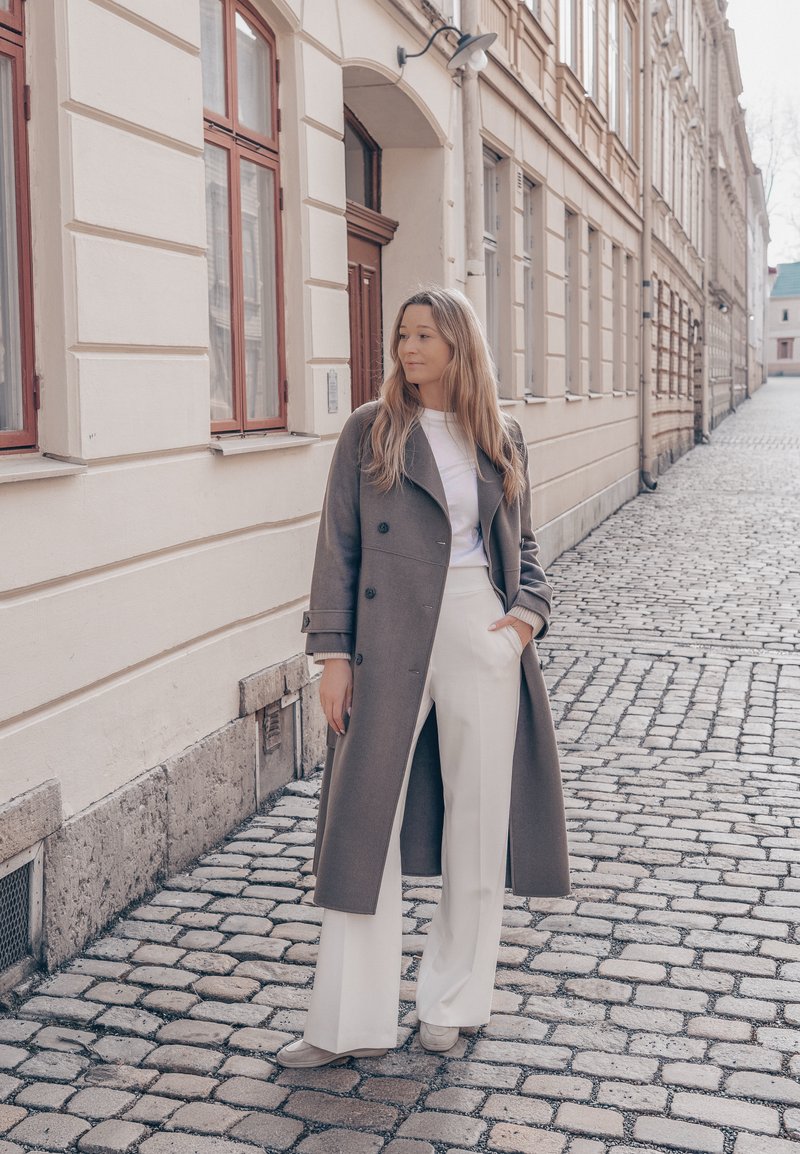 Femme en manteau gris, pantalon blanc et baskets, debout sur une rue pavée à côté de bâtiments beiges avec fenêtres et portes.