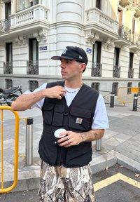 Mladý muž v čiernej čiapke a vestě drží hrnček kávy, upravuje si náhrdelník, stojí na mestskom chodníku s historickými budovami v pozadí.