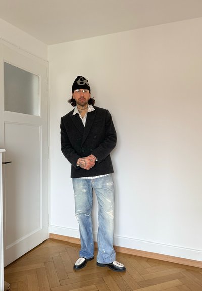 Hombre con tatuajes que lleva gafas, gorro negro, blazer oscuro, camisa a rayas, vaqueros desgastados y zapatos blanco y negro, de pie en el interior sobre un suelo de madera.