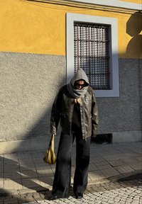 Jaqueta de couro preta, cachecol cinza oversized, suéter preto, jeans largas pretas e uma bolsa verde texturizada. Óculos de sol complementam o visual.