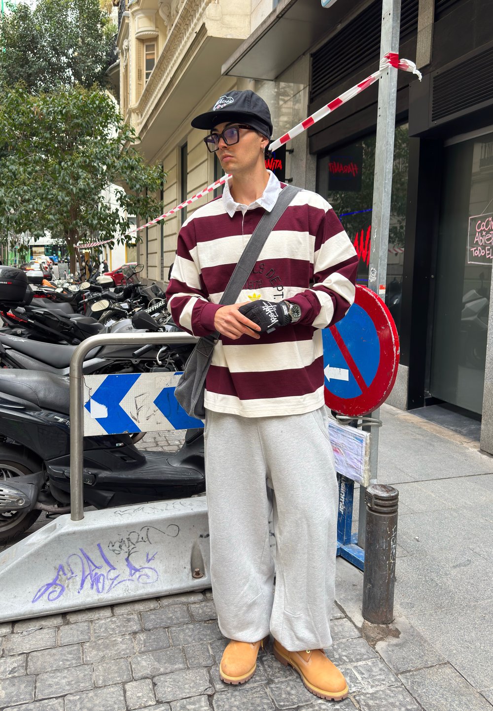 Long-sleeved, maroon and white striped rugby shirt with a collar, grey jogging bottoms, yellow boots, and a black cap. Holds an item in gloved hands.