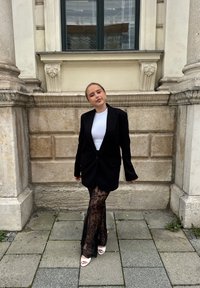 Blazer negro oversize sobre una blusa blanca, combinado con pantalones de encaje negro semitransparente y tacones abiertos blancos, frente a una pared de piedra.